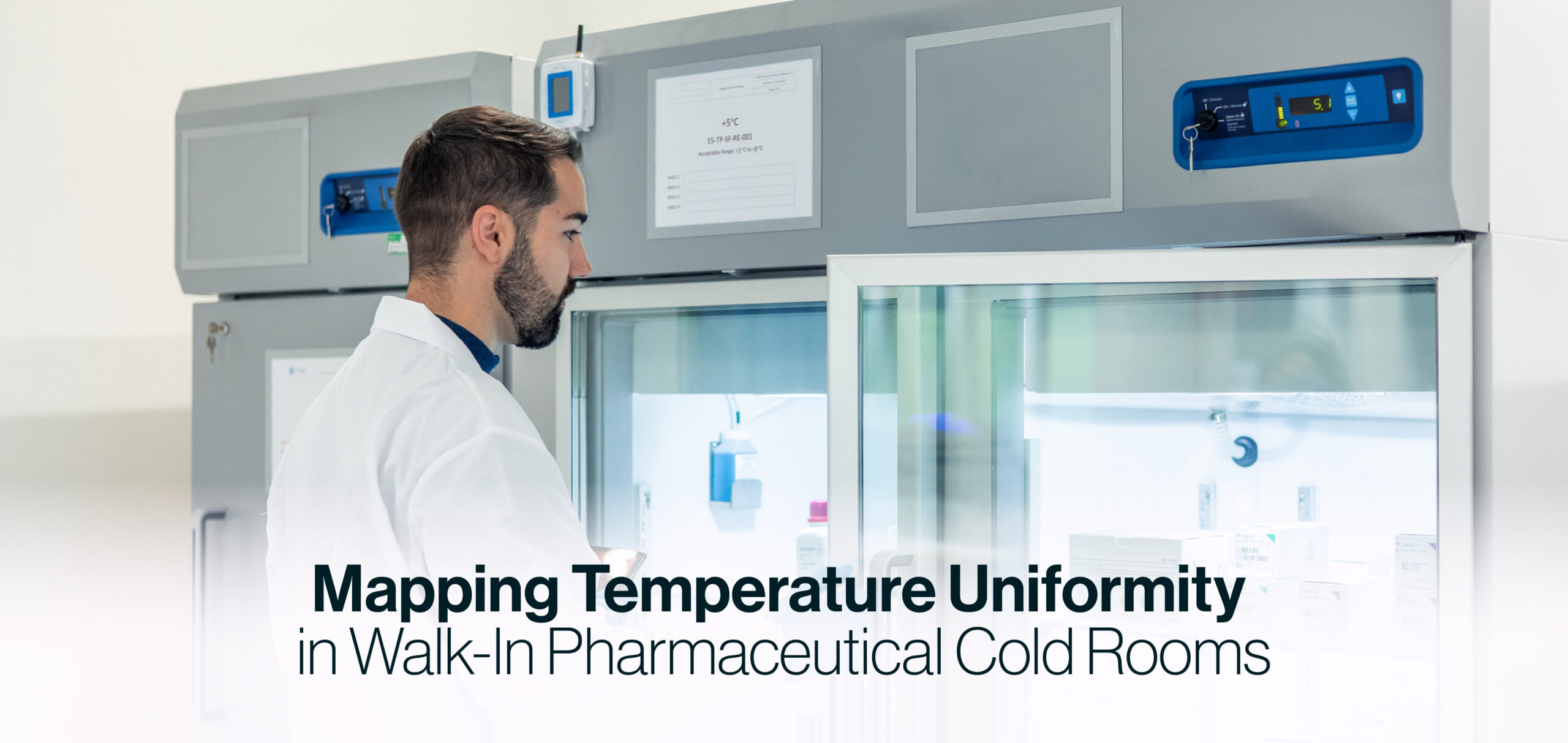 Lab technician in a white coat stands beside a walk-in pharmaceutical cold room, monitoring temperature equipment in use by the facility.