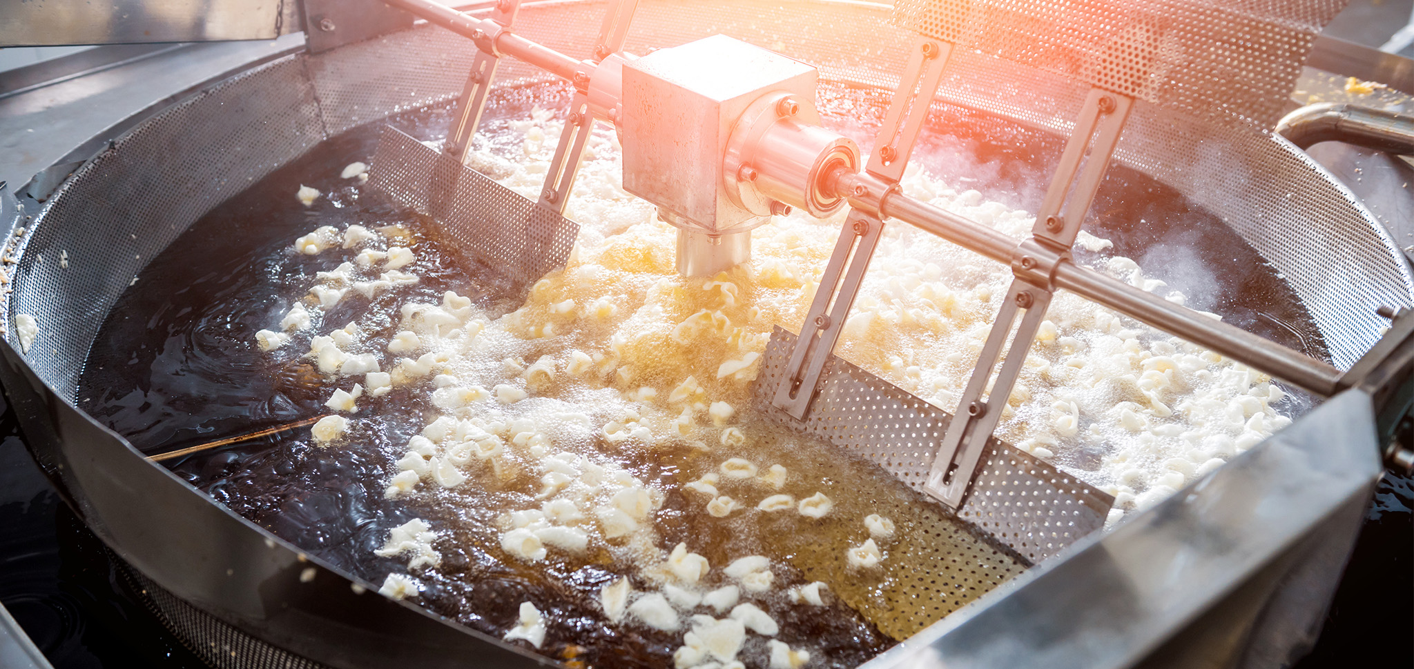 Industrial fryer with perforated metal walls and a rotating arm frying small dumplings in hot oil.